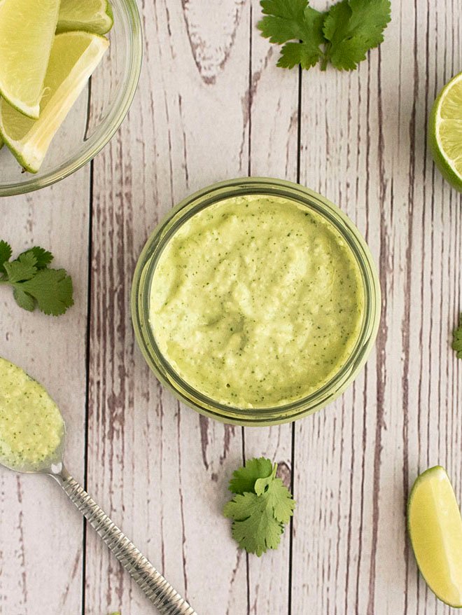 An overhead shot of a small jar of avocado, jalapeno and lime crema, lime slices and sprigs of coriander.