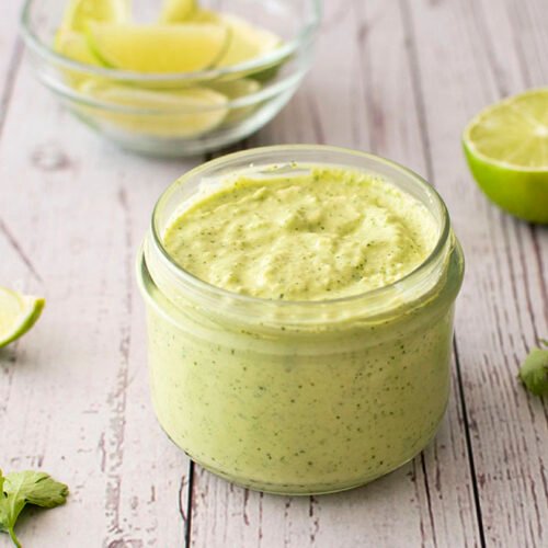 A small jar of creamy avocado, jalapeno and lime crema sitting in front of a small bowl of sliced limes.