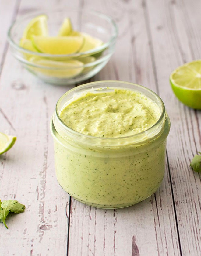 A small jar of creamy avocado, jalapeno and lime crema sitting in front of a small bowl of sliced limes.