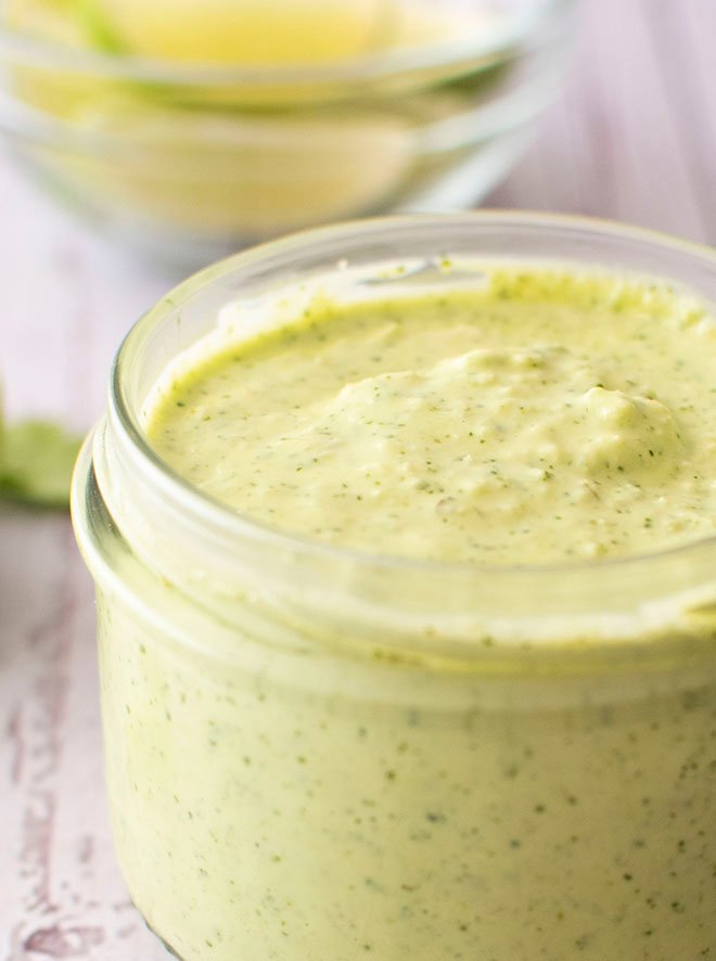 A small jar of creamy avocado, jalapeno and lime crema sitting in front of a small bowl of sliced limes.