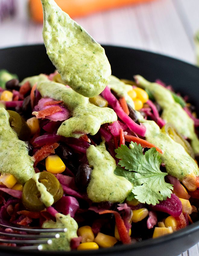 A bowl of Mexican pickled cabbage slaw being drizzled with a teaspoon of avocado, jalapeno and lime crema.