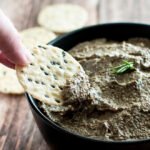 Hand dipping a black sesame and brown rice cracker into the roast mushroom and garlic pate.