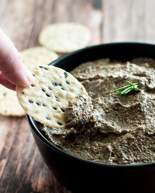 Hand dipping a black sesame and brown rice cracker into the roast mushroom and garlic pate.