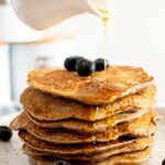 A jug pouring maple syrup on banana buckwheat pancakes,