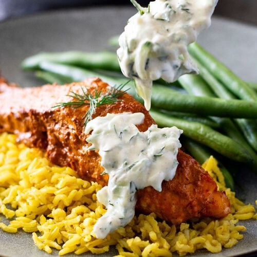 Dill cucumber raita being dripped onto a plate of baked tandoori salmon.