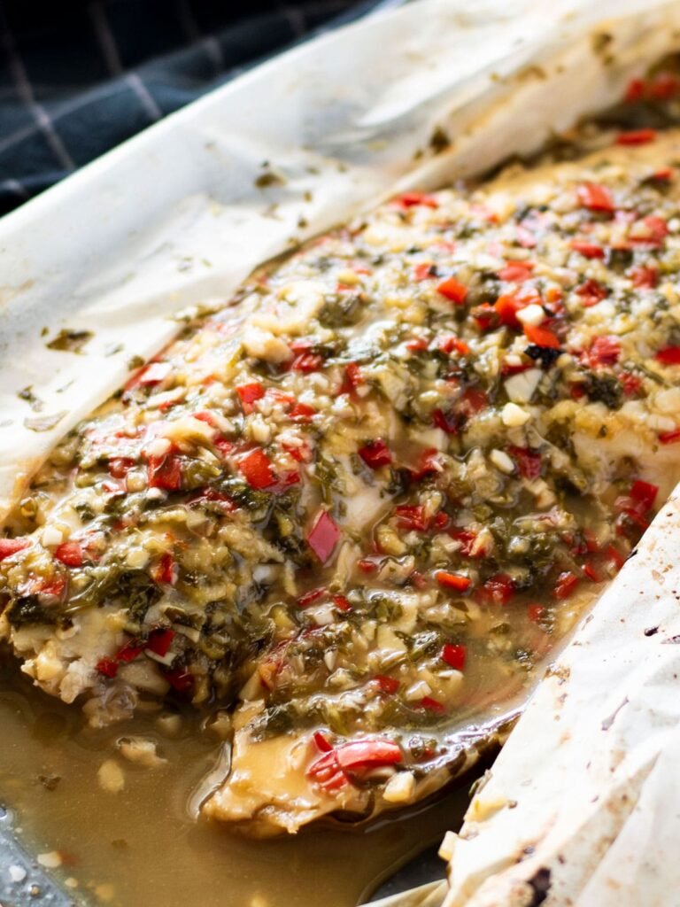 A piece of baked barramundi smothered with chunky Thai sauce inside an opened baking paper parcel.