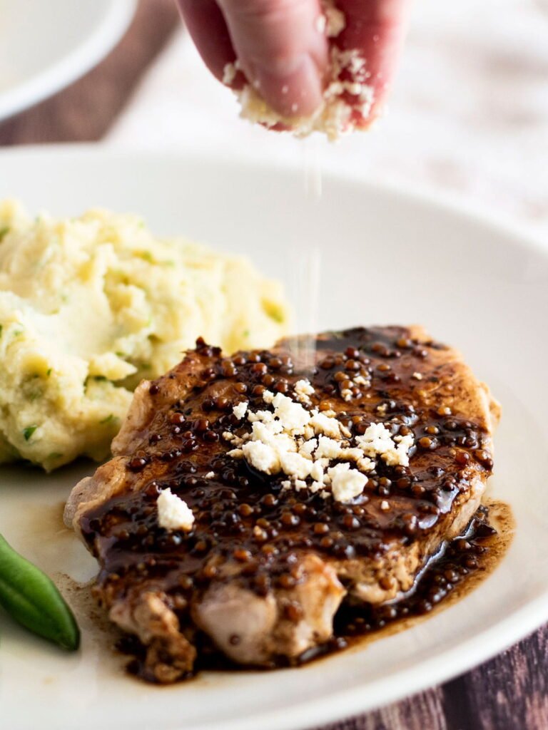 A hand sprinkling fetta cheese onto a pan fried pork steak with balsamic glaze.