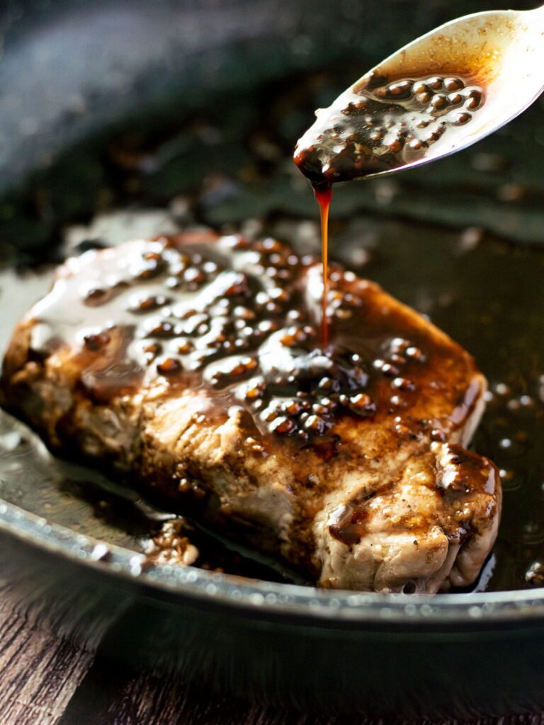 A pan fried pork steak being drizzled with balsamic reduction.