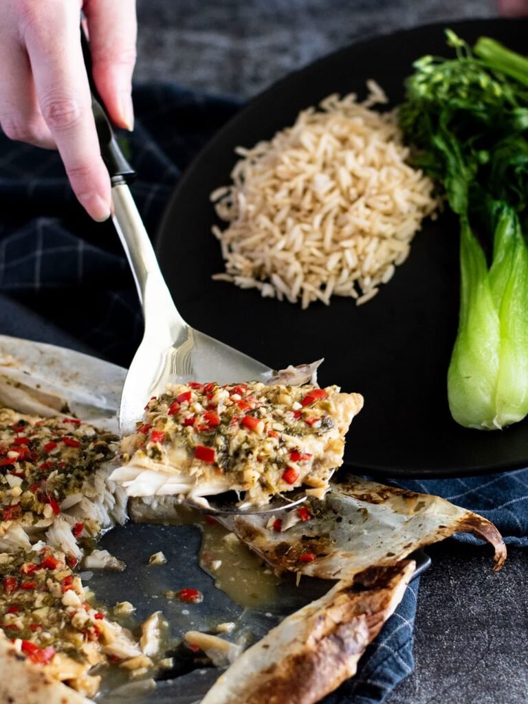 A hand with a ladle scooping a piece of baked barramundi onto a plate with rice and vegetables.