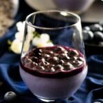 A glass of lavender and blueberry panna cotta with blueberry sauce on navy material.