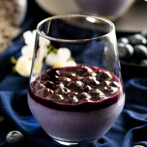 A glass of lavender and blueberry panna cotta with blueberry sauce on navy material.