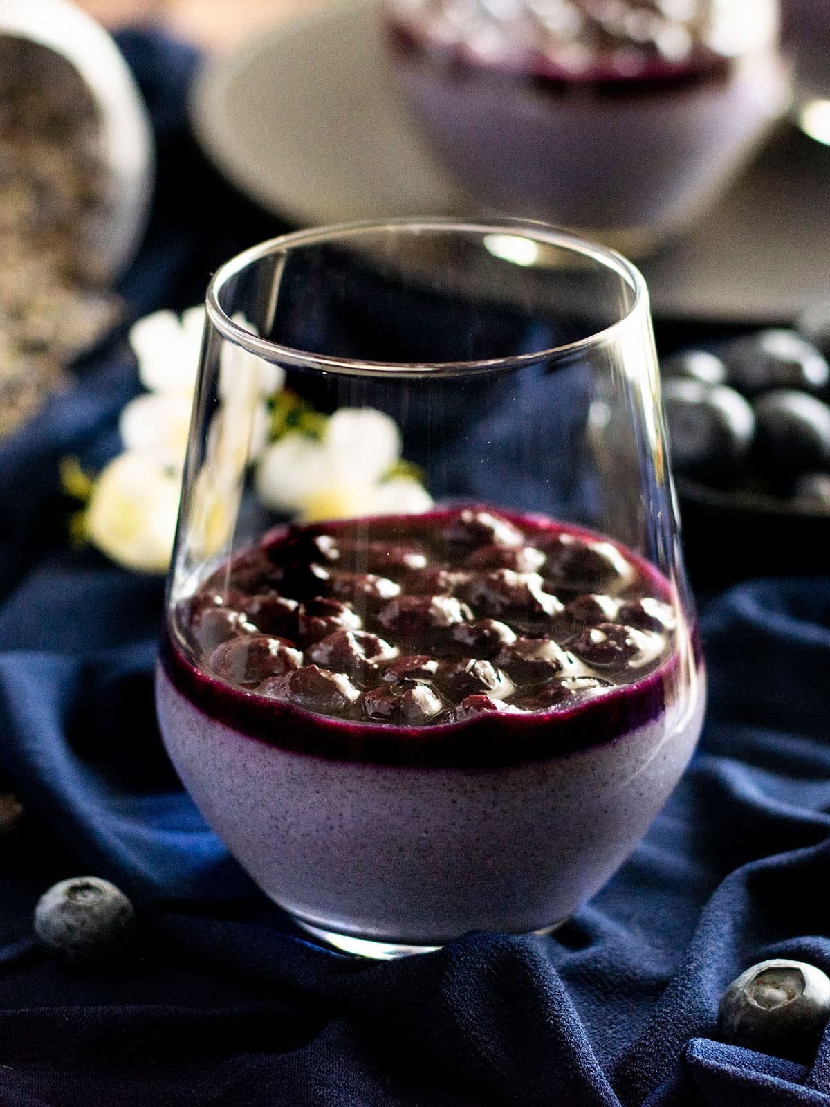 A glass of lavender and blueberry panna cotta with blueberry sauce on navy material.