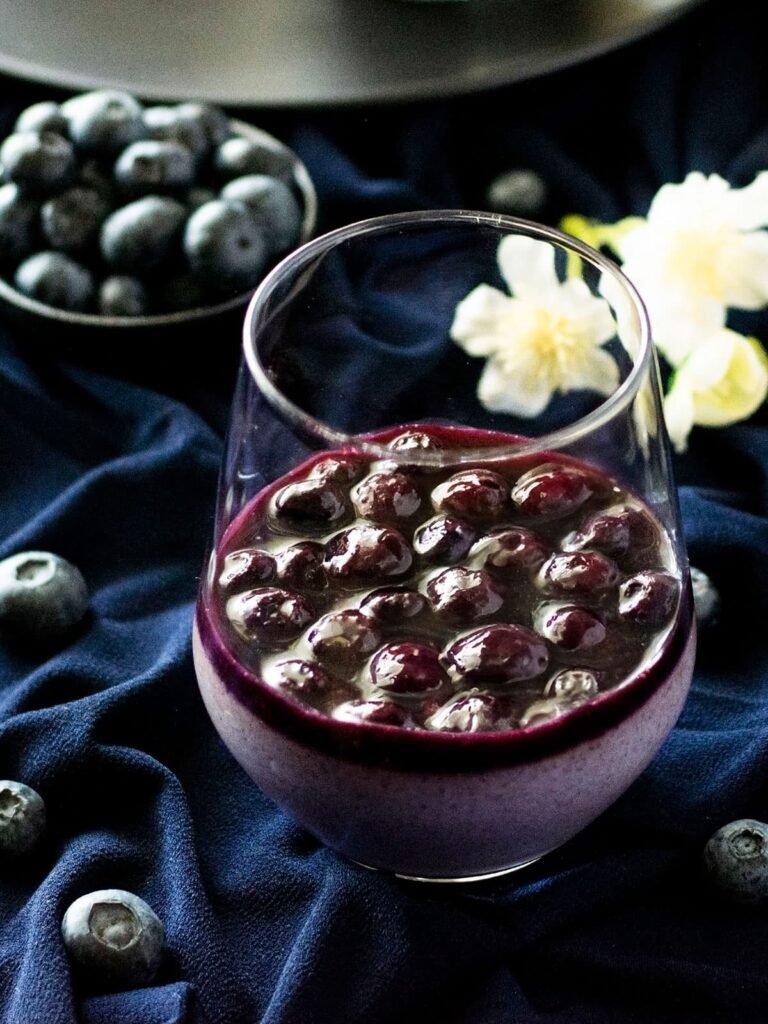 A glass of lavender and blueberry panna cotta on navy fabric.