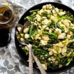 Overhead view of a bowl of warm broccolini salad with spoons and a bottle of dressing.