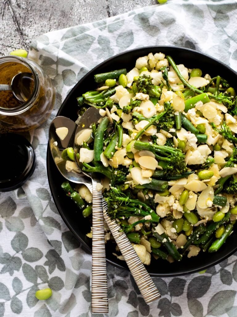 Overhead view of a bowl of warm broccolini salad with spoons and a bottle of dressing.