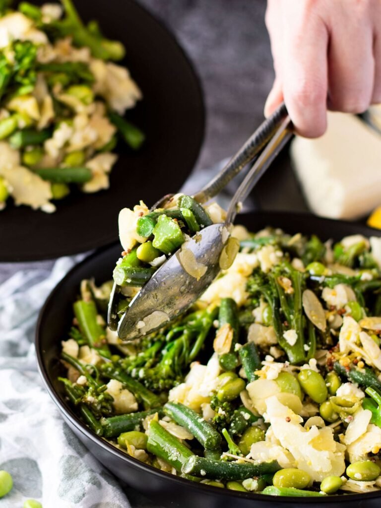 A hand using two spoons to grab some broccolini salad.