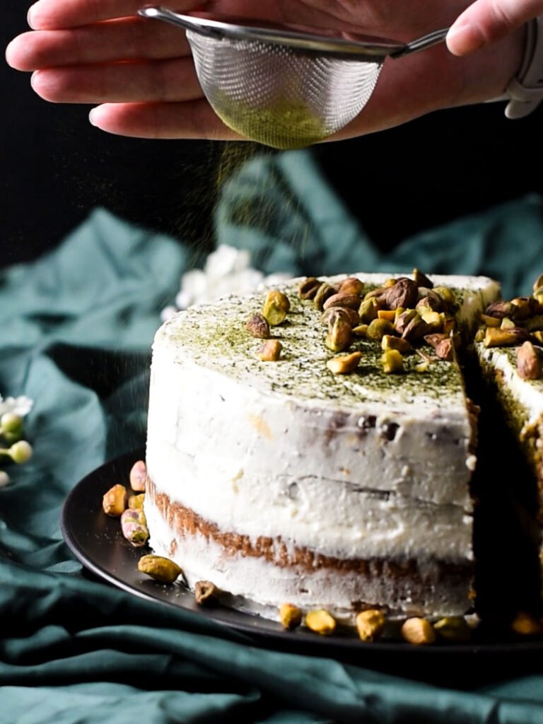 A hand dusting a pistachio and matcha cake with extra matcha.