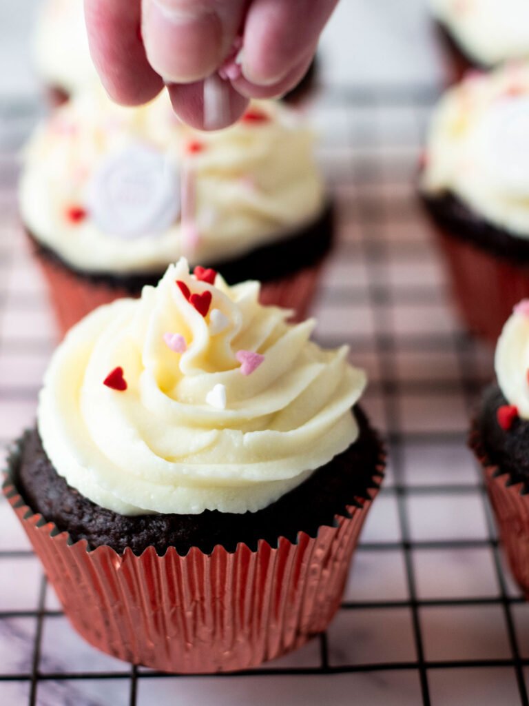 A hand dropping heart sprinkles onto a frosted chocolate beetroot cupcake.