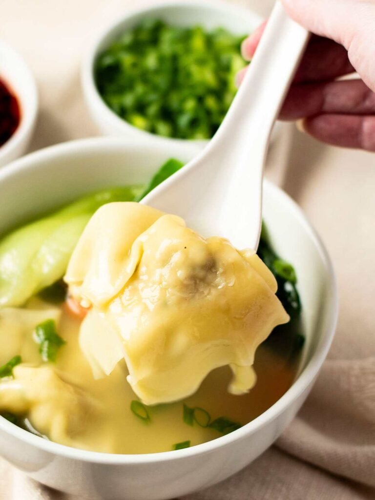 A spoon lifting a wonton out of a bowl of Shanghai wonton soup.
